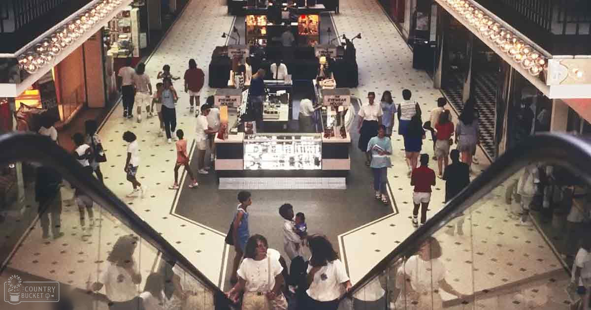 A bustling shopping mall interior from the 1970s-80s, with adults and children in casual attire, with display tables for eyewear under bright overhead lights.