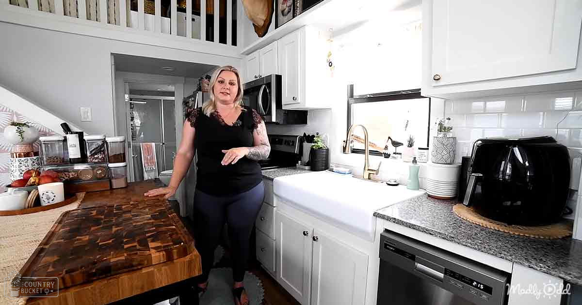 An adult female in a kitchen setting, wearing a black lace-top sleeveless top with dark pants and sandals; various kitchen appliances and decor items are visible.