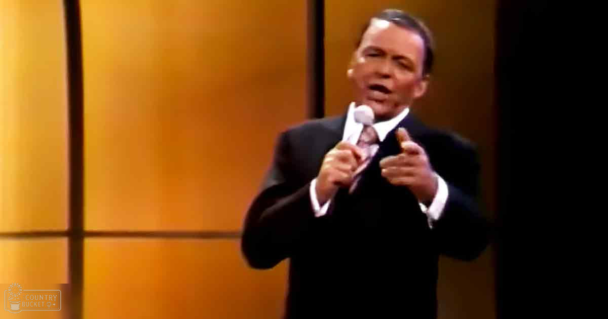 An adult male singer wearing a dark tailored suit jacket, white shirt, patterned tie on a stage with warm-toned vertical lines backdrop.