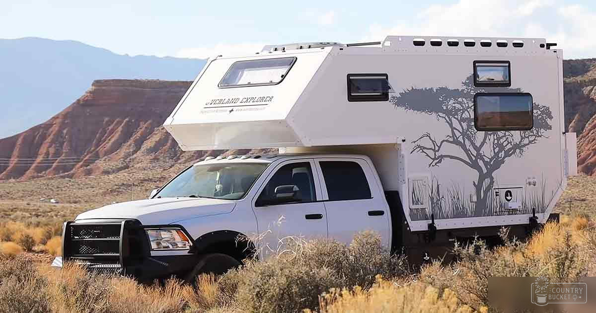 White truck camper with 'Overland Explorer' design in arid desert landscape with red rock formations and low-lying shrubs, partly cloudy sky suggesting midday or early afternoon.