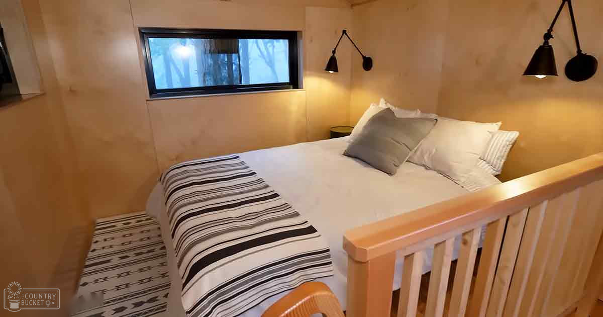 A cozy bedroom scene with a bed, lamps, nightstand, wooden headboard, and patterned rug, natural light, winter or early spring setting.