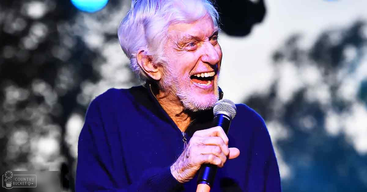 An older adult male singer performing outdoors during daylight, holding a microphone with a blurred green backdrop suggesting a park setting.
