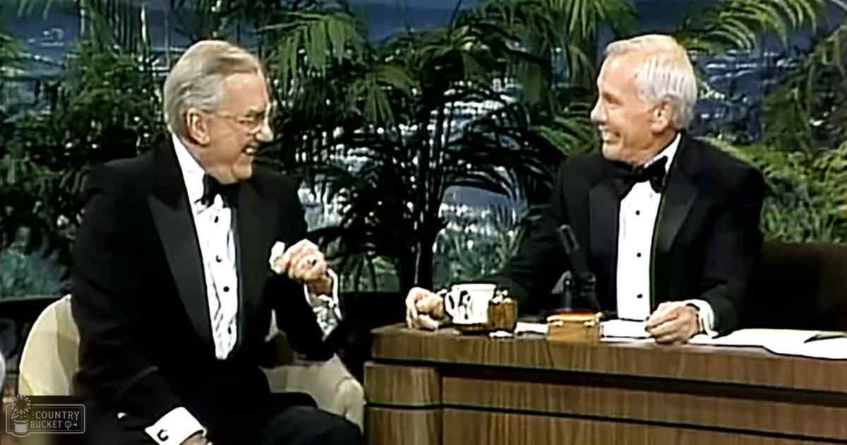 Two older adults dressed in black tuxedos seated on a stage with wooden furniture, decorative elements like plants, and a backdrop suggesting an outdoor cityscape; bright studio lighting.
