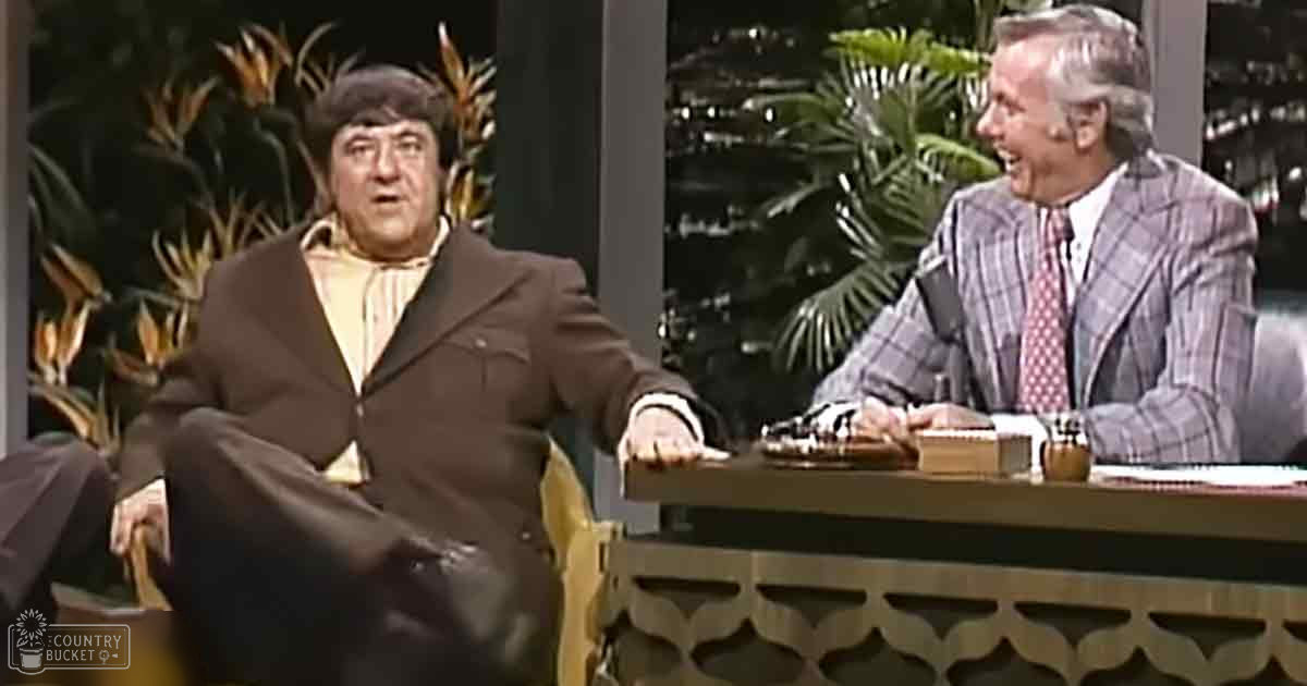 A middle-aged adult male in a brown suit with a yellowish-orange striped shirt sits at a desk with a microphone, while an older adult male in a checkered blazer and red patterned tie is seated to his right. The scene appears to be set on a talk show or interview set.