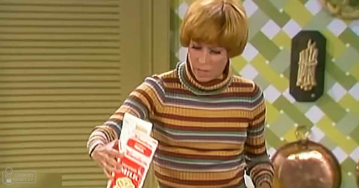 A middle-aged adult female wearing a striped turtleneck sweater indoors, holding a 'Wheat' box with a golden apple-shaped object nearby.
