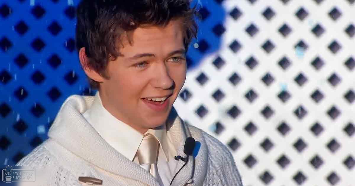 A young adult male wearing a white textured sweater with a hood and a light-colored tie, performing on stage in front of a backdrop with dark squares.