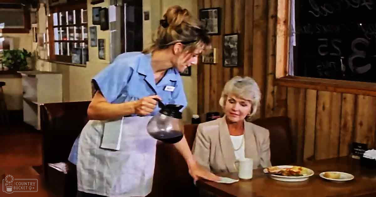 A young adult female waitress in a diner-style restaurant wearing a blue uniform top and white apron serves coffee while an older adult woman, dressed in a beige blazer over a white top with a pearl necklace, sits at a table eating food.