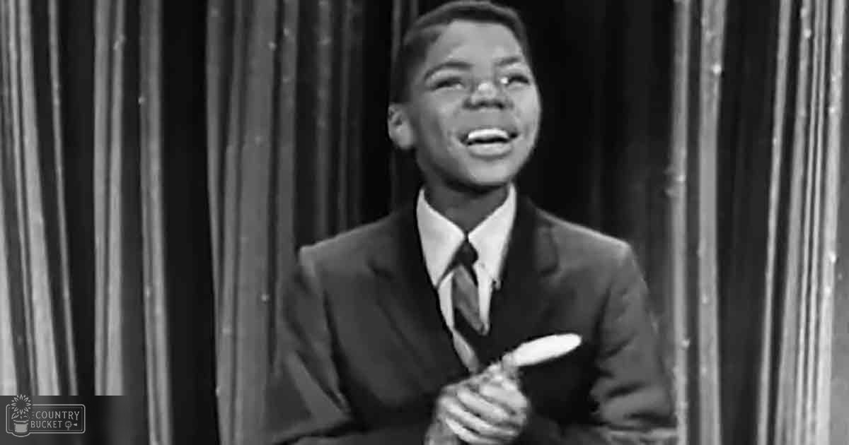 Young adult male in a formal suit holding a microphone, wearing 'Madly Odd' text on his jacket; monochromatic setting with curtains and stage-like backdrop.