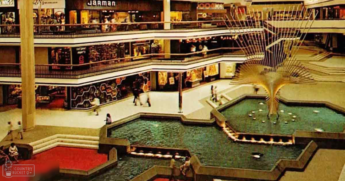 A shopping mall interior with a central decorative fountain and multiple levels of stores. Children and adults are present, wearing casual attire.