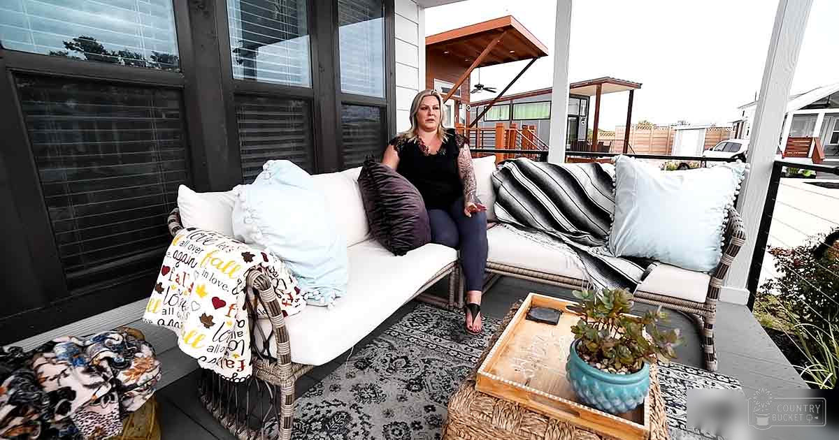 A middle-aged woman sits in an outdoor seating area of a modern home, surrounded by wicker furniture and decorative pillows with 'fall' text.