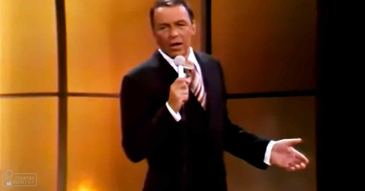 An adult male performer wearing a dark, mid-20th-century style suit with white shirt and patterned tie on stage with warm-toned panels in the background.
