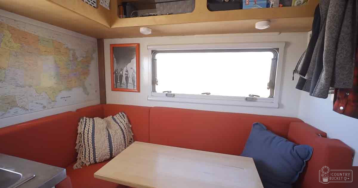 Compact RV or camper van interior with red sofa, wooden table, map of North America on wall, framed picture above window, and various clothing items hanging.