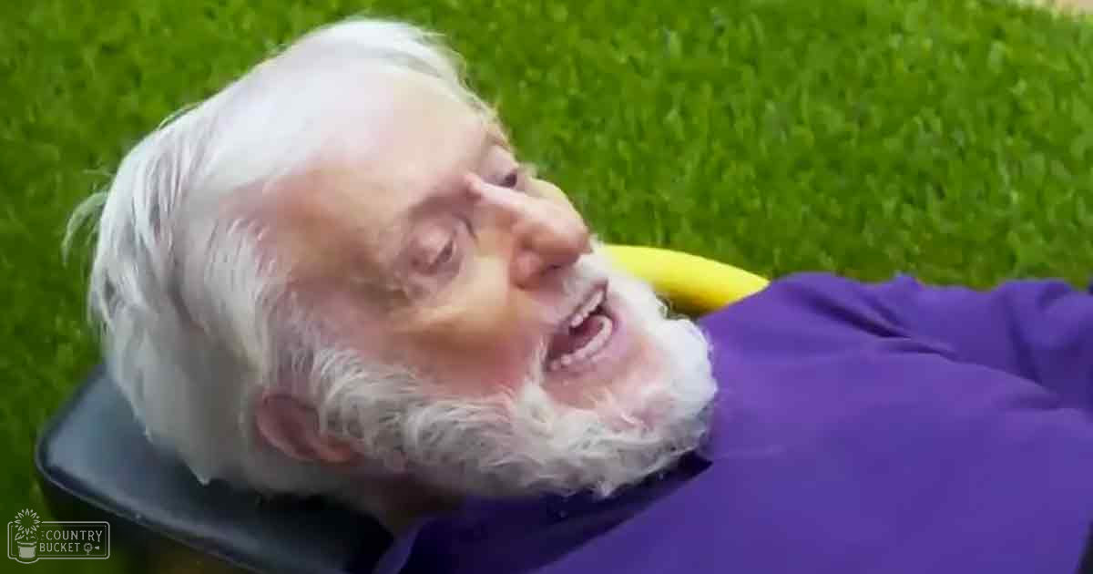 An older adult male with white hair and a beard, wearing a purple tunic-style robe on a green grassy outdoor setting.