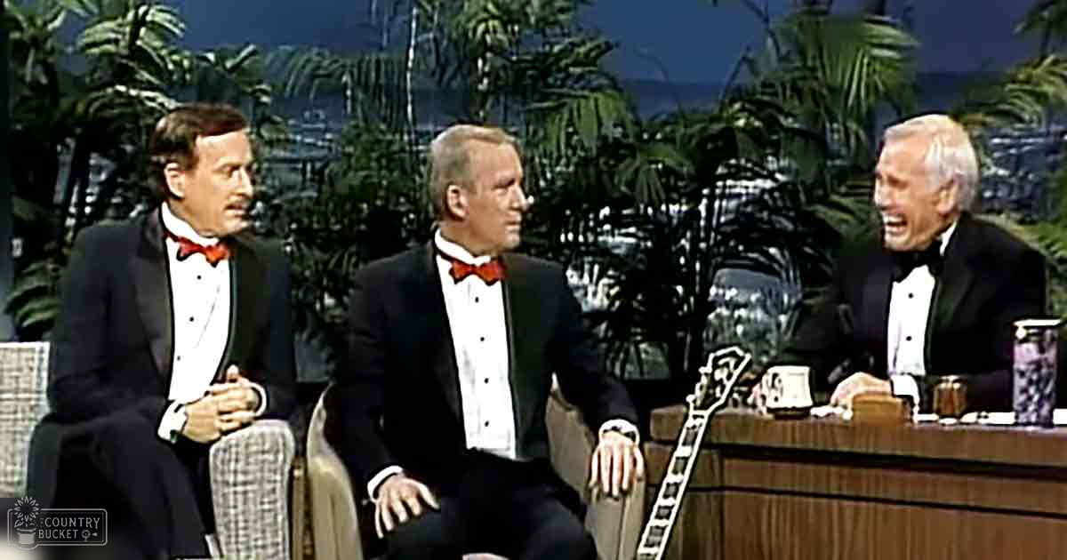Talk show setting with three adult males in black tuxedos, palm tree backdrop, and studio lighting typical of the 1960s-1970s.