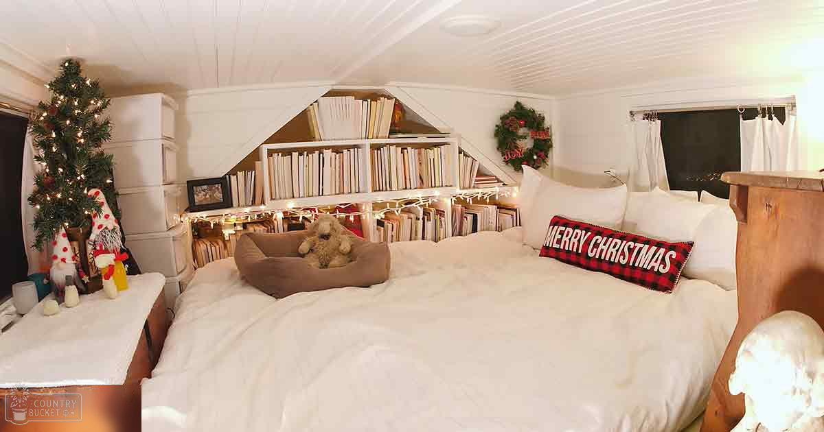 A stuffed dog toy rests in a cozy Christmas-decorated bedroom with white bedding, wooden headboard, and various holiday decorations.