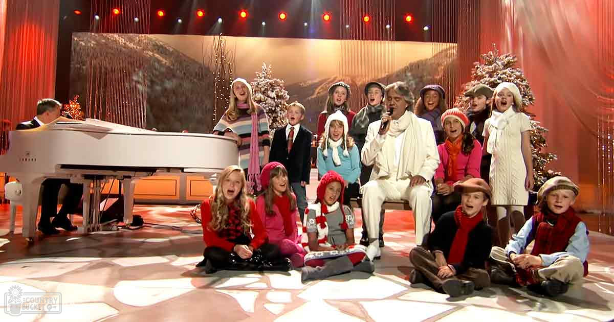 A group performance scene in a studio setting with adults and children dressed for winter, with a white grand piano and Christmas trees.