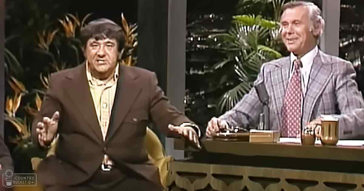 Two adult males seated at a talk show set; one wears a dark brown suit jacket with a patterned tie from the mid-20th century, while the other sports a plaid blazer and red patterned tie. The setting includes a wooden desk adorned with plants and papers, suggesting an indoor studio environment.