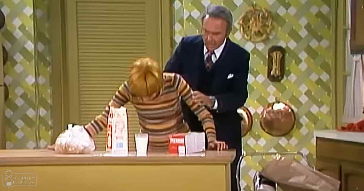 A toddler girl in a striped shirt with an apron, wearing light-colored pants, indoors near a wooden counter with milk cartons and food items.