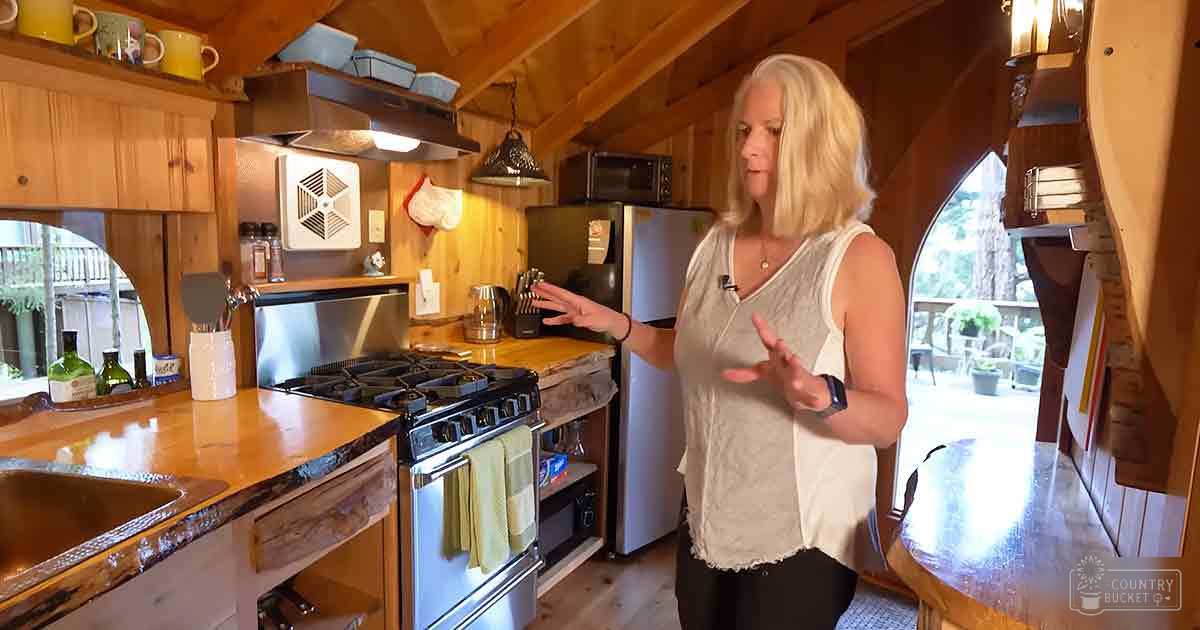 A middle-aged adult female wearing a sleeveless, light gray, round-neck top with a frayed hem and accessories in a rustic kitchen setting with wooden cabinets, stainless steel appliances, and natural daylight.