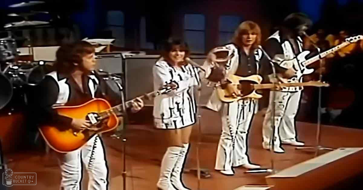 A musical performance with young adult female performers in a stage setting holding acoustic guitars; an adult male singer with electric guitar, dressed in white pants and dark vest.