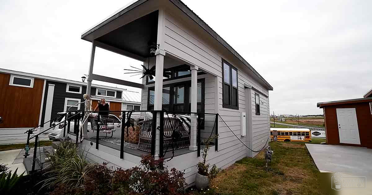 A modern tiny home with a porch in an outdoor residential area, with a person sitting on the swing. The setting suggests spring or summer with grassy surroundings.