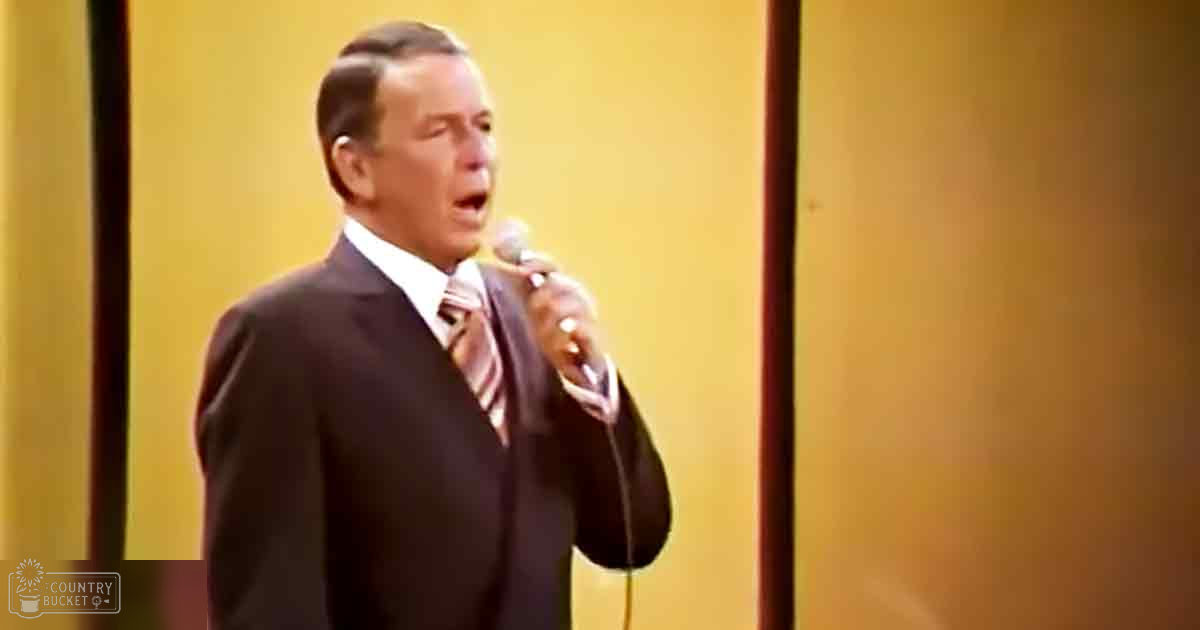 A middle-aged man wearing a dark suit with a striped tie sings into a microphone against a yellow backdrop.