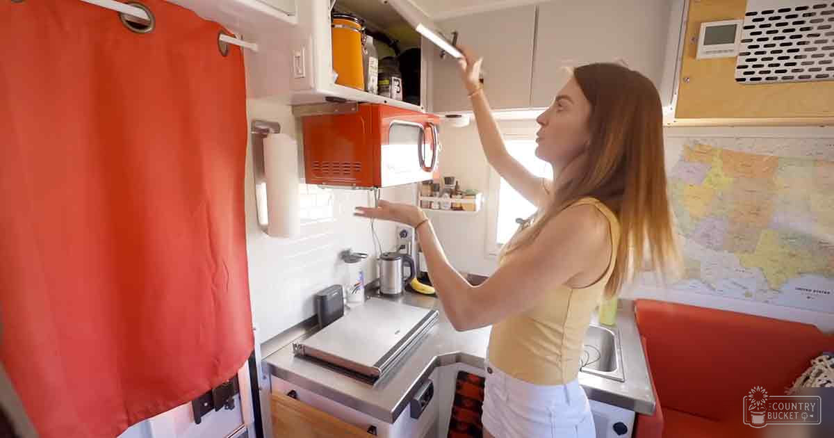 A young adult female in a compact RV kitchen with natural light streaming through. A red curtain hangs on the left side, while a small kitchenette with appliances is visible.