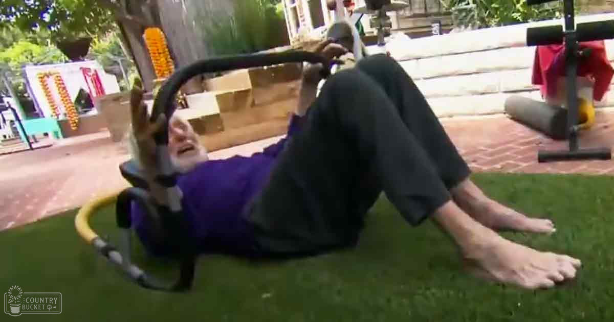A middle-aged adult male lies on a green artificial turf lawn, holding onto a green hose while wearing a purple shirt and black pants. The scene includes an outdoor setting with a white fence, tree trunk decorated in orange, and colorful elements suggesting a festive or play area.