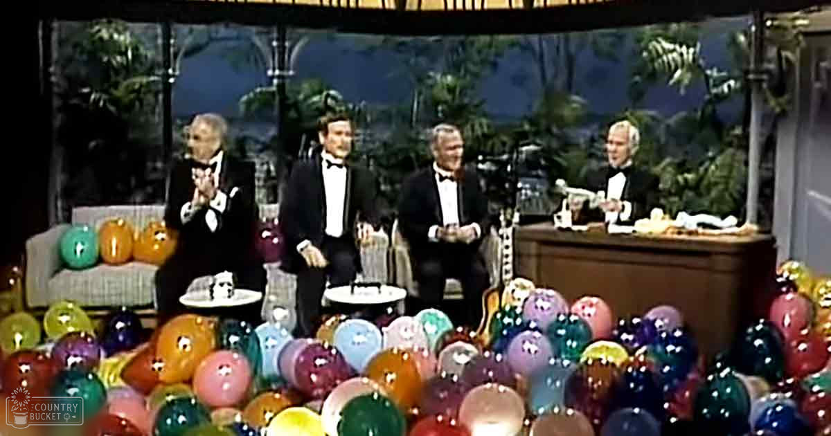 A comedic skit in an indoor studio with a tropical backdrop with palm trees through large windows. The setting includes a couch, pumpkins on tables, and a desk resembling a judge's bench.