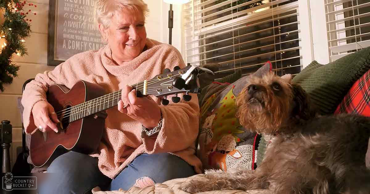 A middle-aged adult female playing guitar in her home with a Brussels Griffon dog, surrounded by holiday decorations and natural daylight.