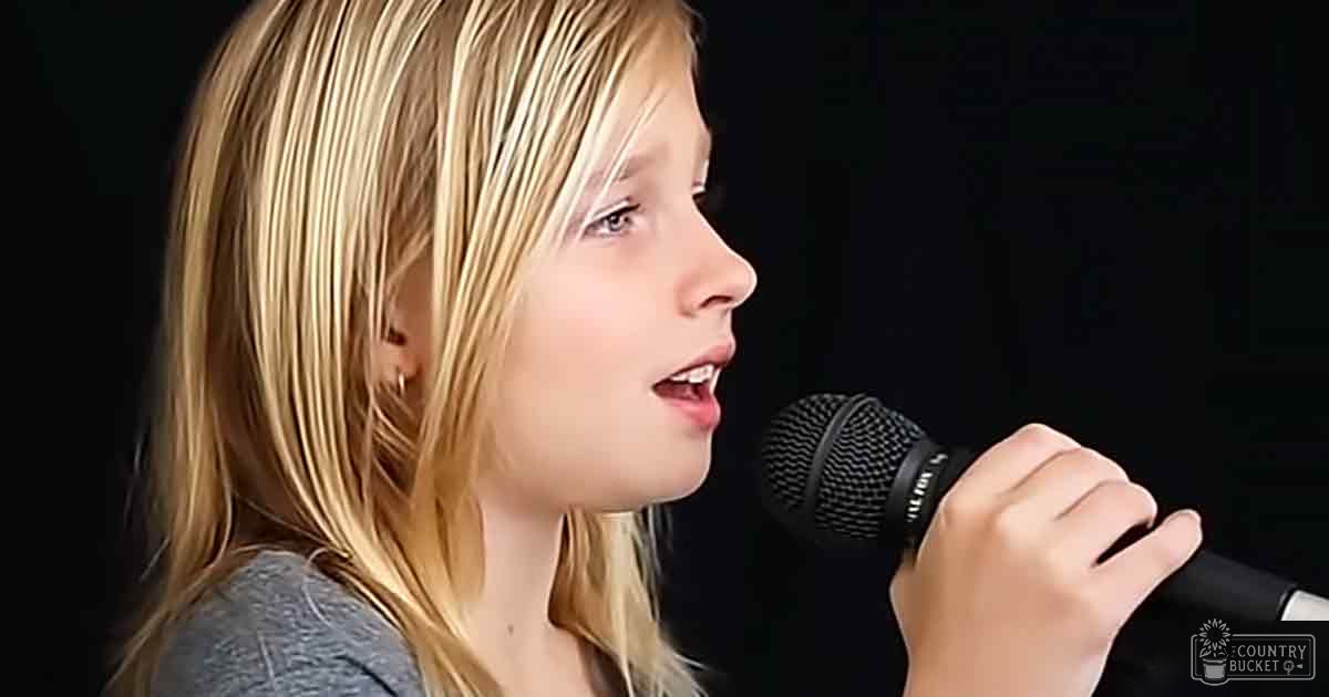 A young adult female sings into a microphone, wearing a gray long-sleeved crew neck top indoors.