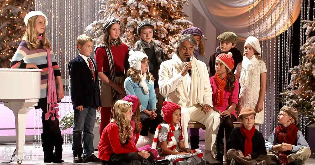 An adult male singer performing on stage with children dressed for a holiday event; Christmas trees decorated with lights and ornaments visible in the background.