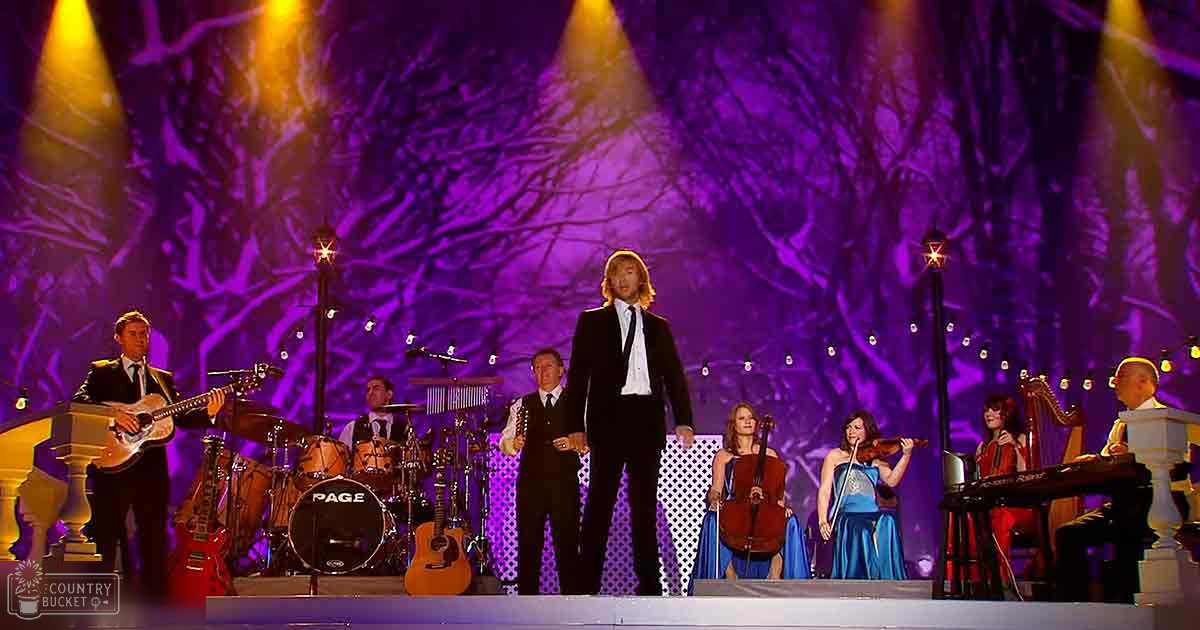 Musical performance in an indoor concert setting with a stage backdrop with tree-like branches and warm stage lighting at night.