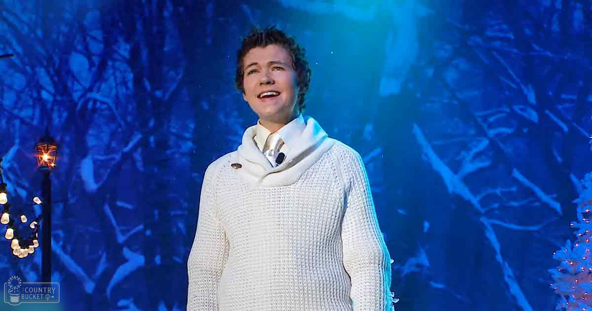 A young adult male in a performance setting, wearing a relaxed, modern white knitted sweater with high collar and long sleeves against silhouetted trees under blue stage lighting.