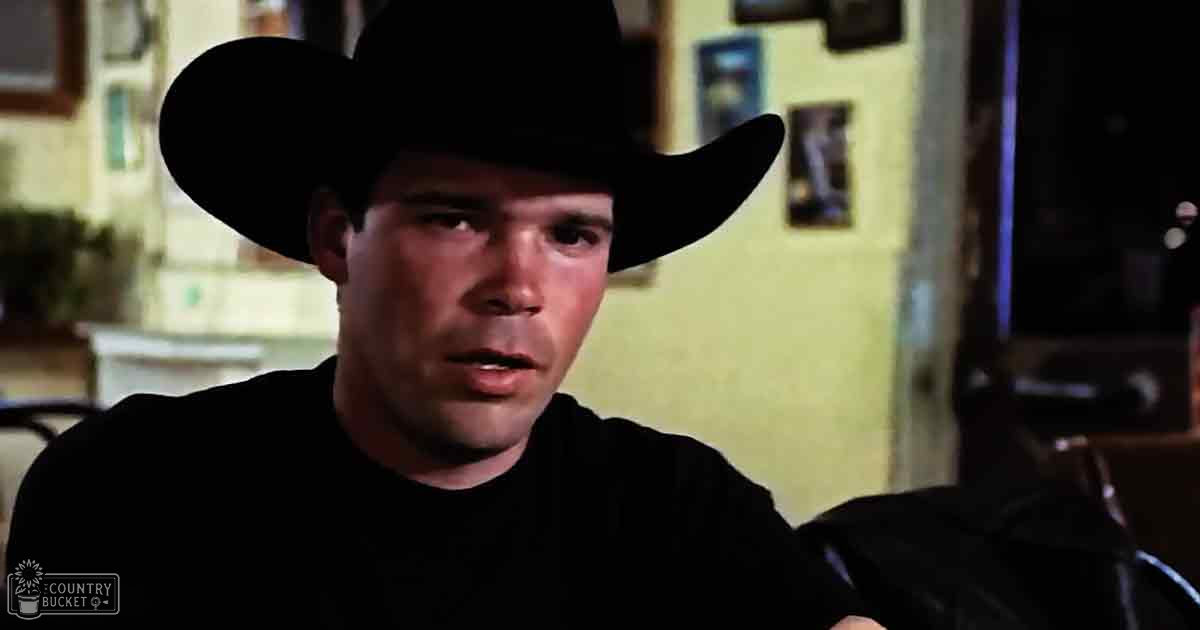 Young adult male wearing a black cowboy hat, simple black t-shirt, indoor setting with artificial lighting.