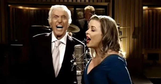 A duet singing into microphones in a mid-20th century recording studio with dark-toned attire and professional equipment.