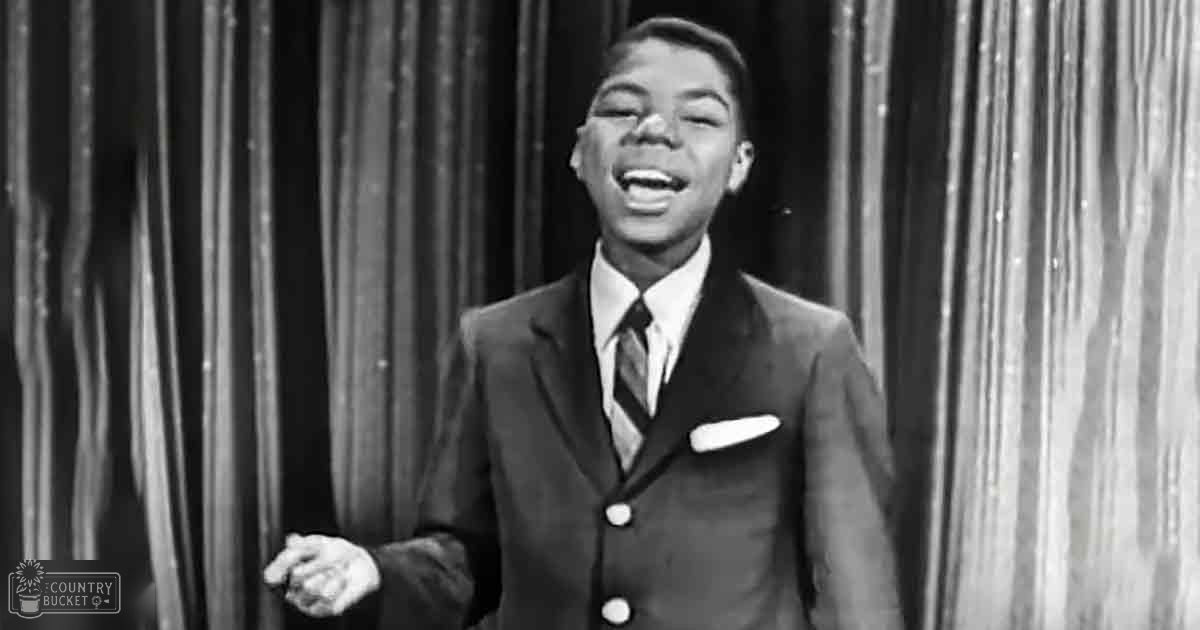 A young adult male in a tailored three-piece suit with a white pocket square, standing on stage with curtains behind him.