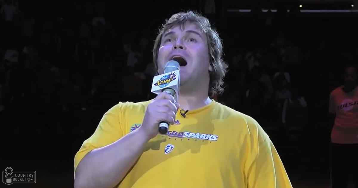 A young adult male singer wearing a yellow 'Los Angeles Sparks' T-shirt with an Adidas logo, holding a microphone in front of a blurred sports arena setting.