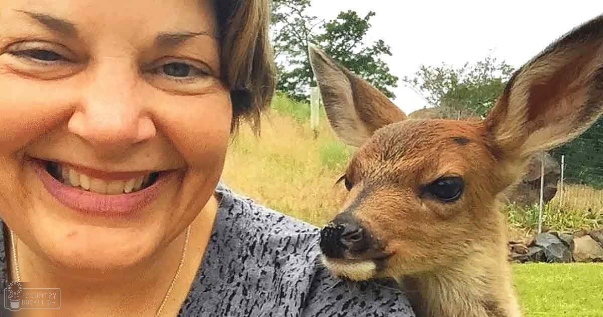 A middle-aged woman wearing a patterned top in an outdoor rural setting with a juvenile deer nearby.