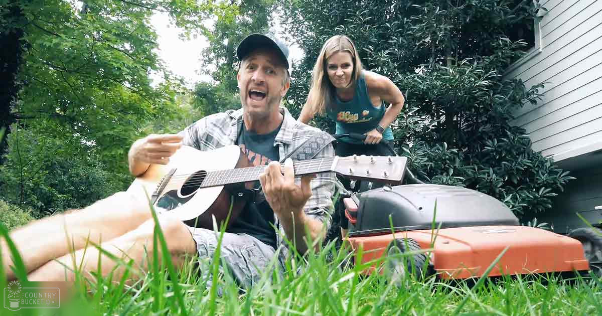 Middle-aged man playing guitar outdoors amidst greenery with a lawn mower visible.