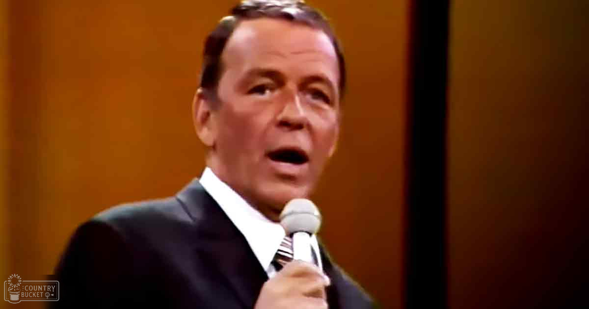 A middle-aged man wearing a dark suit jacket and striped tie holds a microphone against an indoor backdrop with warm stage lighting, suggesting a performance setting.