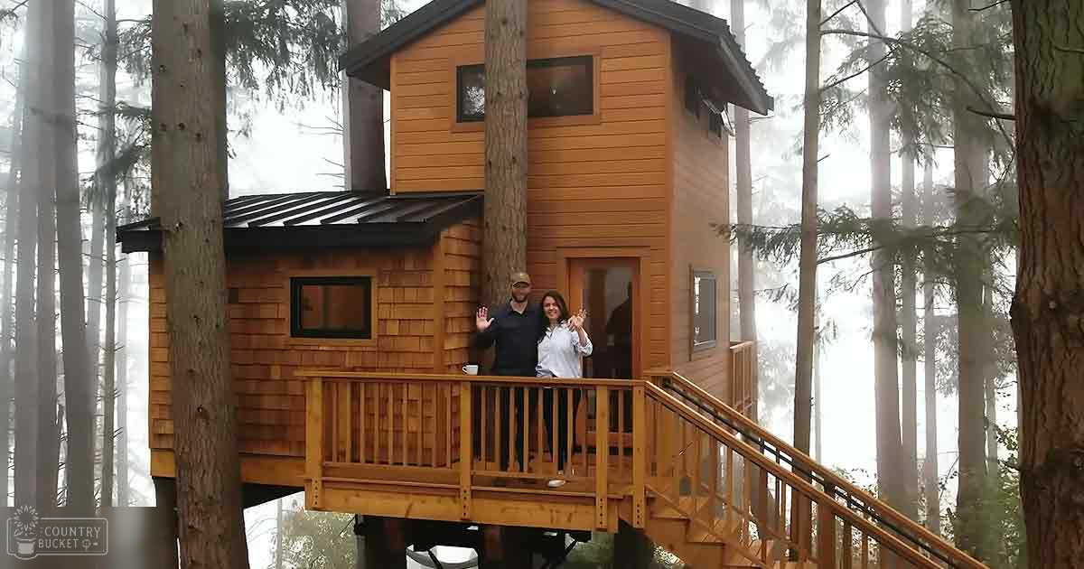 A couple stands on a rustic treehouse porch surrounded by tall forest trees under an overcast sky.