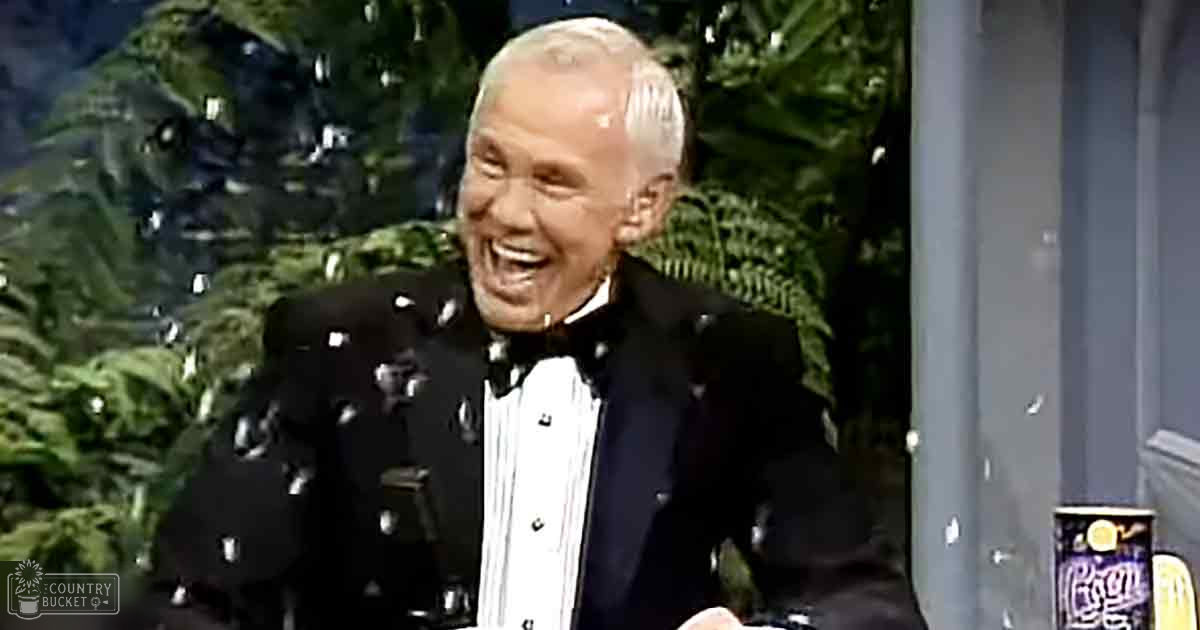 A middle-aged adult male in a classic formal black tuxedo with a bow tie sits indoors amidst lush greenery and artificial lighting.