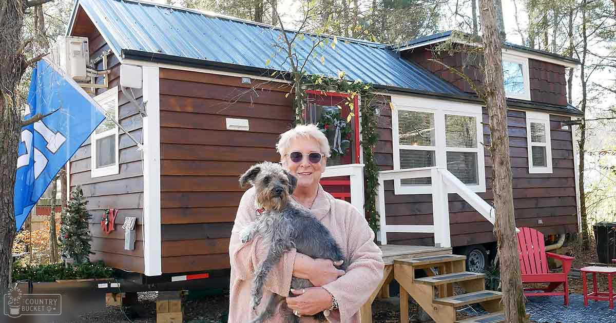 An older adult female holding a gray terrier mix dog in front of a red Adirondack chair with Christmas decorations on a tiny house amidst trees, suggesting late fall or early winter.