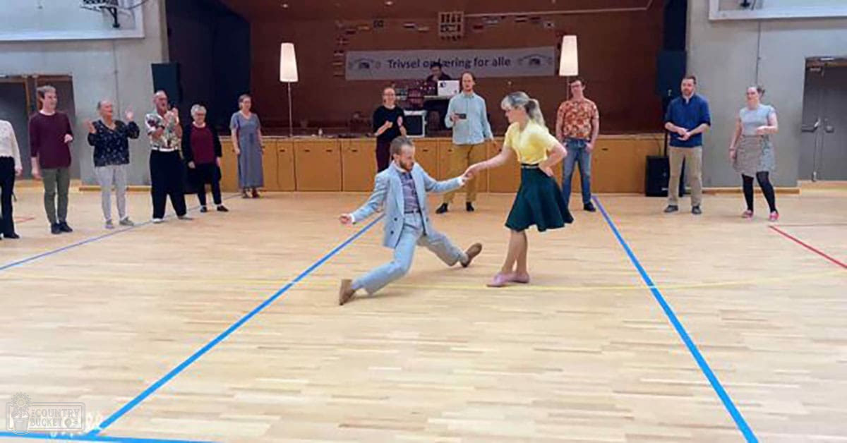 Two adults dancing in a community center with onlookers, wooden floors marked for dance practice, natural light streaming through windows.