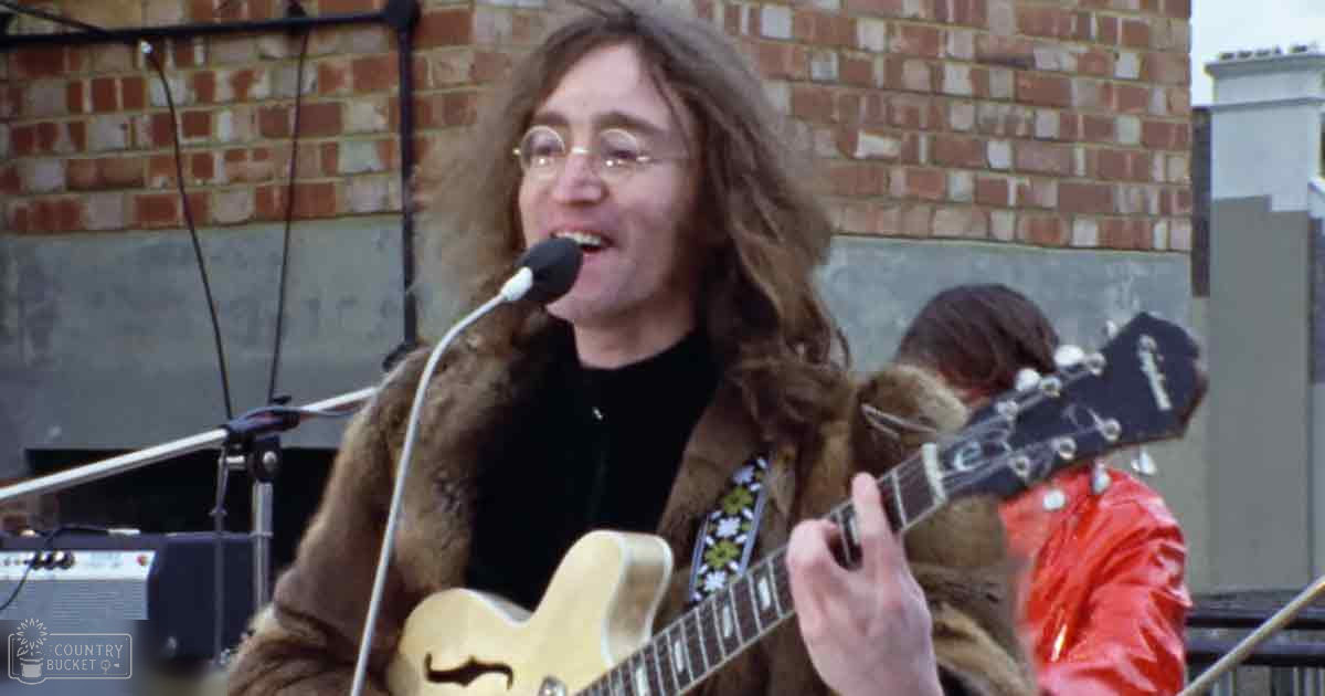 Man playing acoustic guitar outdoors, wearing brown floral-patterned fur coat and black shirt, round glasses, brick wall urban setting, spring or summer daytime natural light.