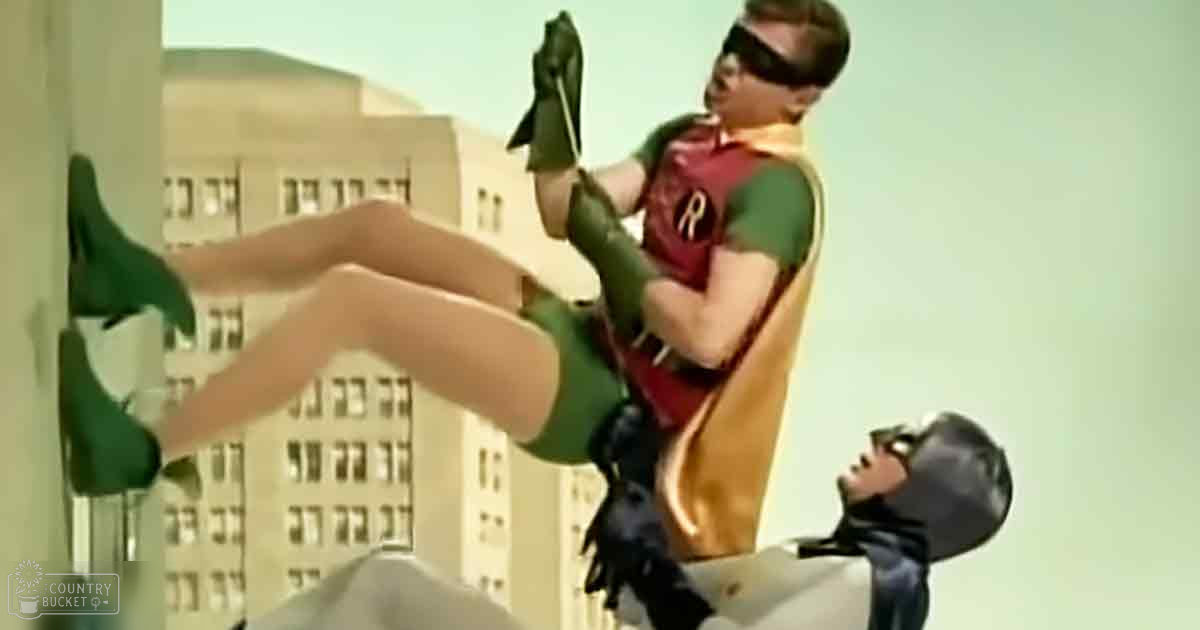 A superhero character in a red and green costume stands atop tall buildings against a backdrop of spring or summer light, with another person in a white shirt and black mask below. The urban setting is sunlit with clear skies.