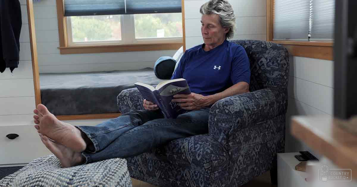 A middle-aged adult female reading in a home setting with white paneling walls, natural light from windows, and casual indoor furniture.