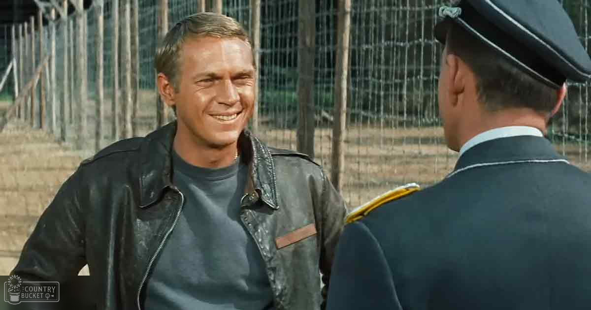 A young adult male wearing a dark gray t-shirt under a black leather jacket with brown accents stands in an outdoor setting, possibly a prison yard. An older adult male in a dark blue uniform with gold epaulettes and a peaked cap is seen in the background.