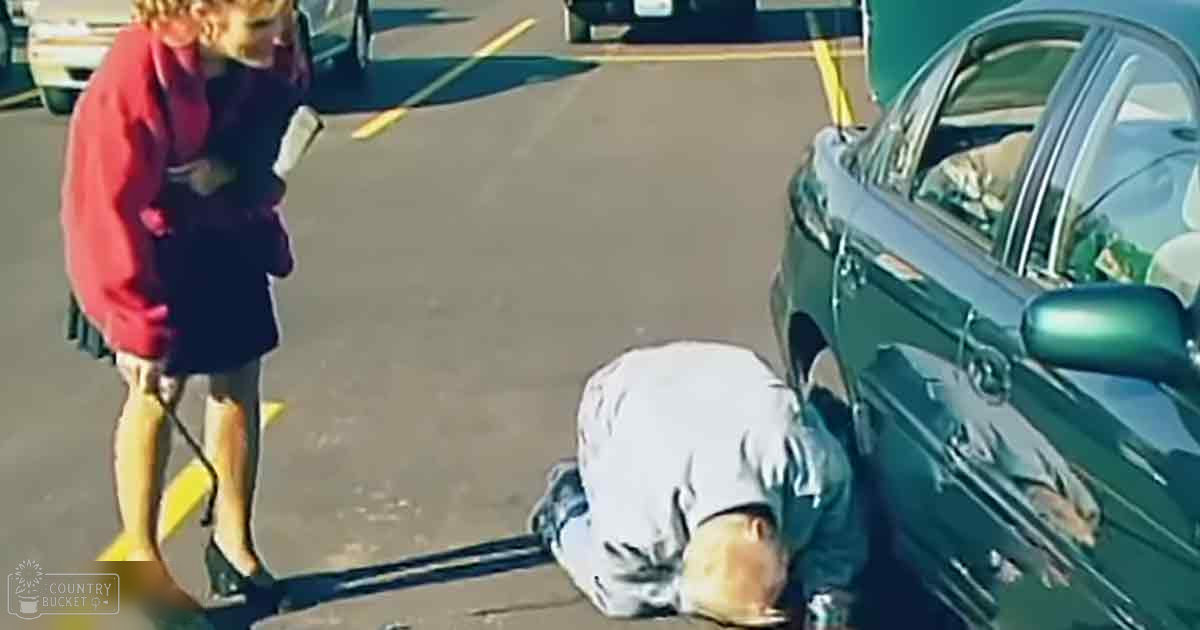 An adult male lying face down in front of a green sedan with another female standing nearby, both outdoors in what appears to be a spring or summer parking lot setting.